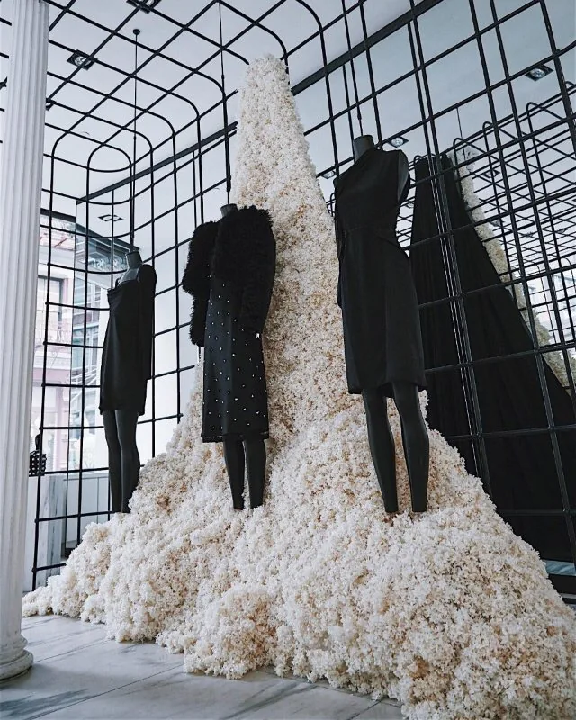 Escaparate de campaña con maniquíes vestidos de negro sobre una montaña de flores blancas y una estructura de rejilla negra