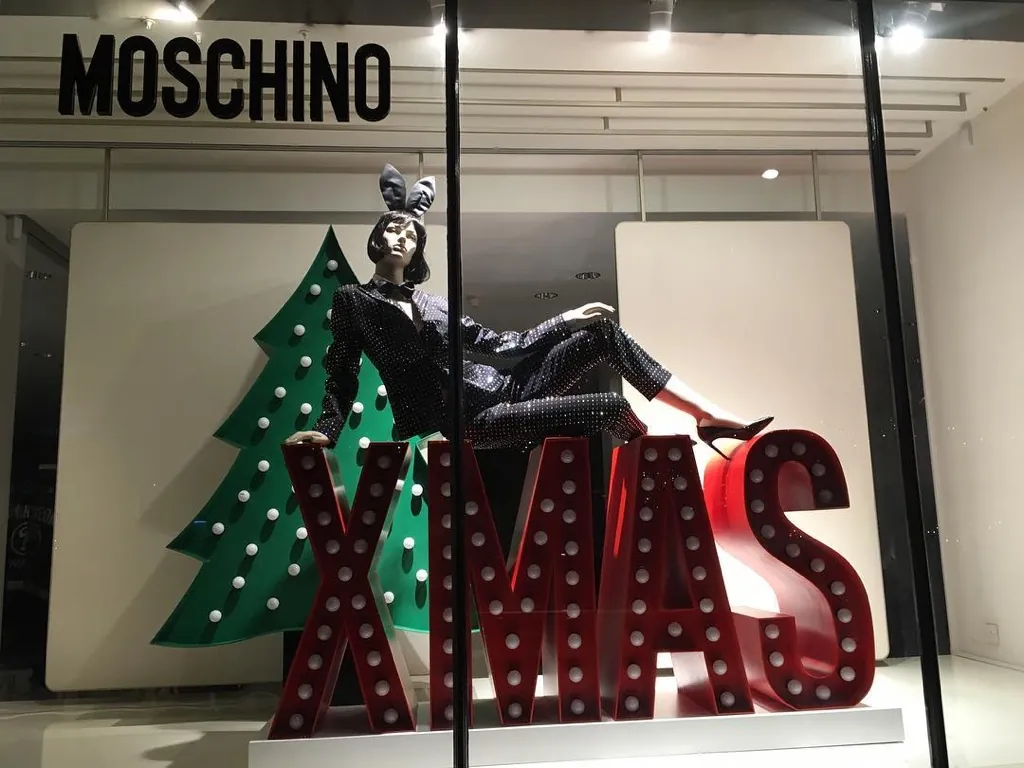 Escaparate de Navidad con letras XMAS rojas iluminadas con bombillas, un árbol verde y un maniquí recostado encima