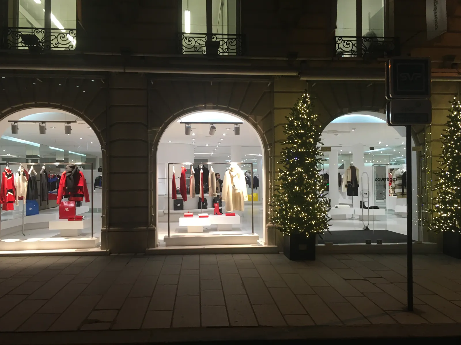 Fachada de una tienda de moda de noche, con escaparates en arco iluminados y árboles de Navidad junto a la entrada