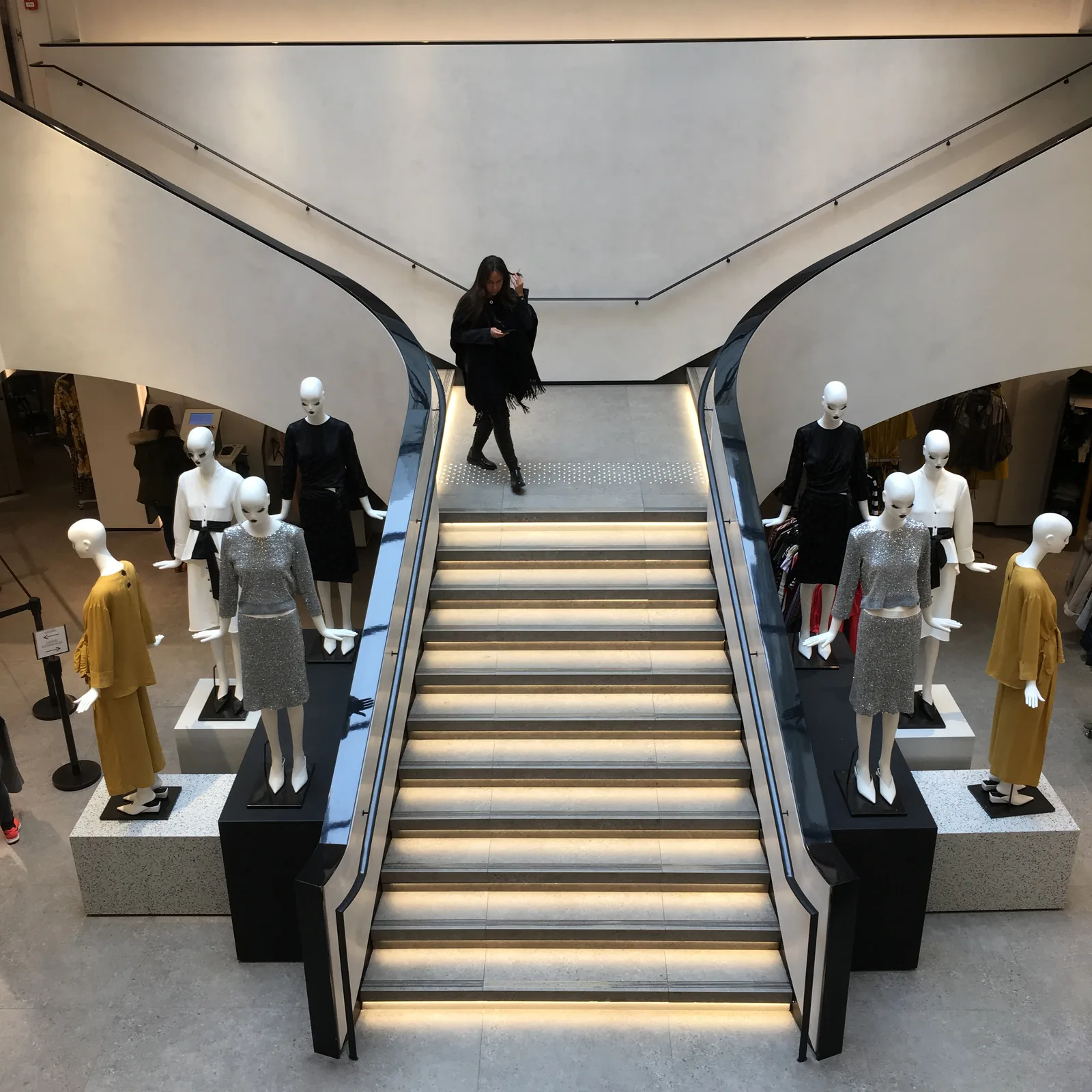 Interior de tienda de varias plantas con escalera central iluminada y grupos de maniquíes a ambos lados